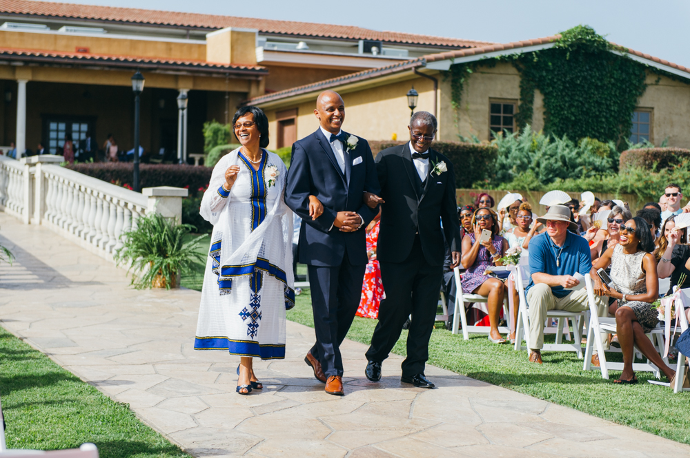 Groom walks down the aisle with his parents on either arm. They are all smiling as seated wedding guests look on with smiles.

Luxury Texas Wedding Photographer. Timeless Wedding Photography. Wedding in Texas. Destination Wedding Photographer.