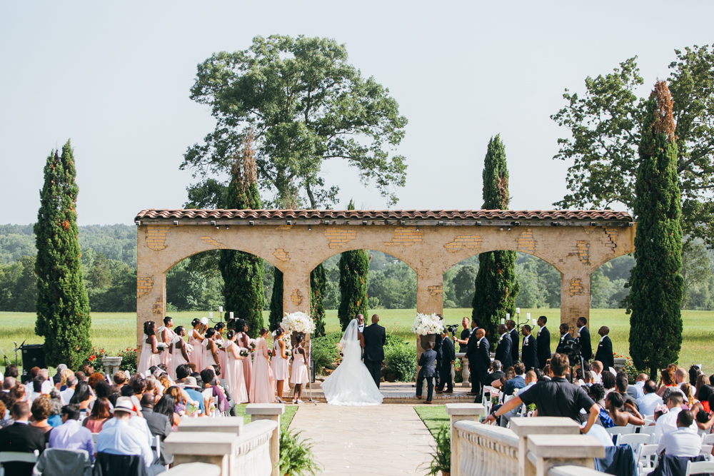 Wide shot of the entire wedding ceremony. Wedding guests are seated, the bride and groom stand at the altar with the bridal party at their side. There is a tri-archway behind them as well as a grassy field and trees.

Luxury Texas Wedding Photographer. Timeless Wedding Photography. Wedding in Texas. Destination Wedding Photographer.