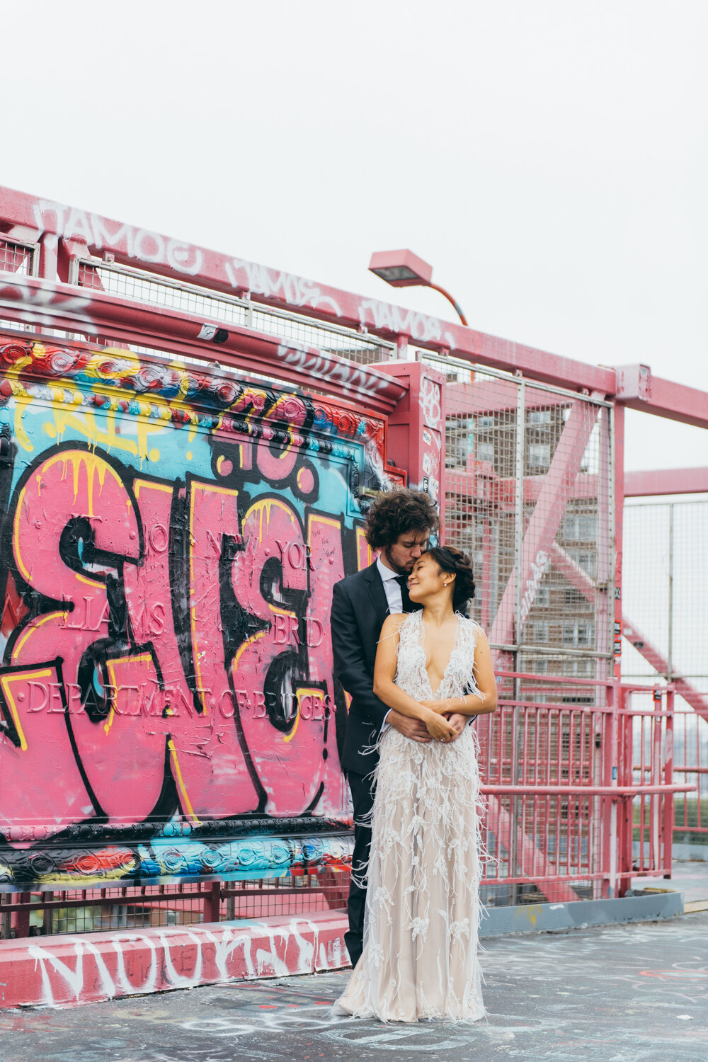 Bride and groom stands on the Williamsburg Bride in front of a wall of graffiti. He is standing behind her with arms around her and he kisses her forehead as she smiles with her eyes closed.

Central Park Wedding Photography. Williamsburg Bridge Bridal Portraits. Luxury NYC Wedding Photographer. Manhattan Micro Wedding.