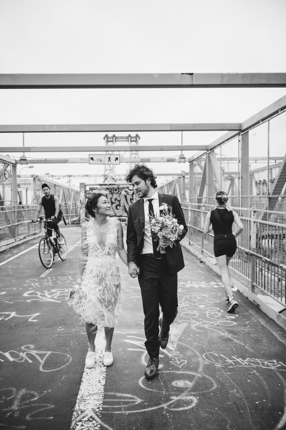 Bride and groom walk along the Williamsburg Bridge holding hands. The groom is holding the bride's flower bouquet as she is holding up the skirt of her feathered wedding dress in her hand. Runners and bicyclists pass them on the sides.

Central Park Wedding Photography. Williamsburg Bridge Bridal Portraits. Luxury NYC Wedding Photographer. Manhattan Micro Wedding.