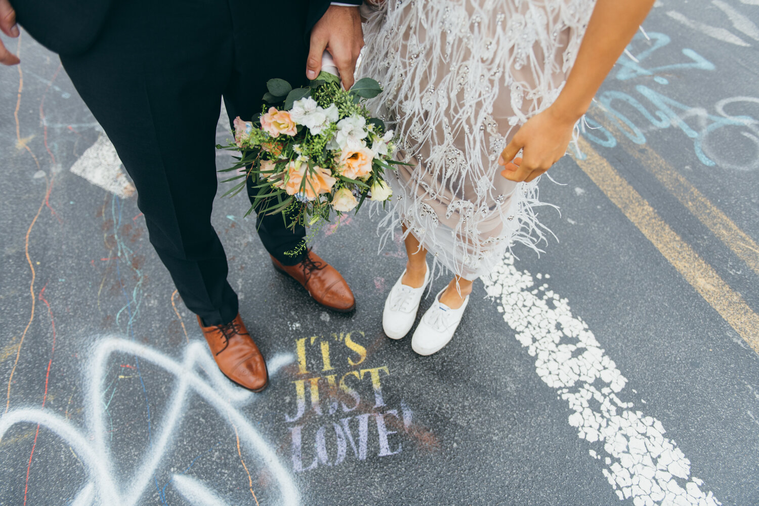 Bride and groom's feet and legs are photographed from above. The bride is holding up the skirt of her feathered dress to reveal white sneakers. They are standing beside a graffiti stencil on the pavement that says "IT'S JUST LOVE"

Central Park Wedding Photography. Williamsburg Bridge Bridal Portraits. Luxury NYC Wedding Photographer. Manhattan Micro Wedding.