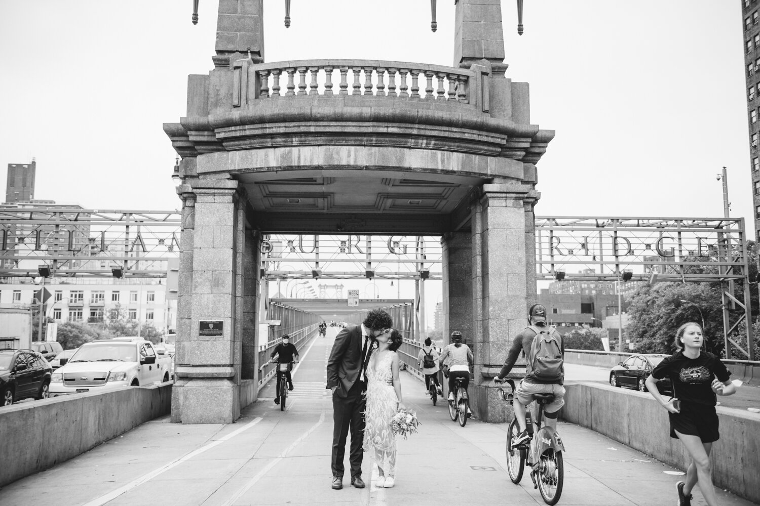 Bride and groom stand at the entrance to the Williamsburg Bridge and kiss below the sign that says "WILLIAMSBURG BRIDGE"Bicyclists and runners pass them on each side.

Central Park Wedding Photography. Williamsburg Bridge Bridal Portraits. Luxury NYC Wedding Photographer. Manhattan Micro Wedding.