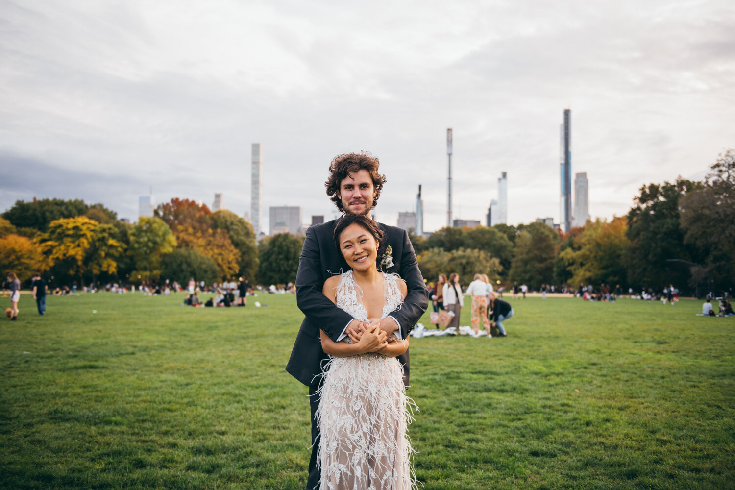 Groom stands behind the bride and has his arms around her. They both smile at the camera. They are standing on the grass in Central Park and park goers, trees, and Manhattan buildings are visible in the background.

Central Park Wedding Photography. Williamsburg Bridge Bridal Portraits. Luxury NYC Wedding Photographer. Manhattan Micro Wedding.