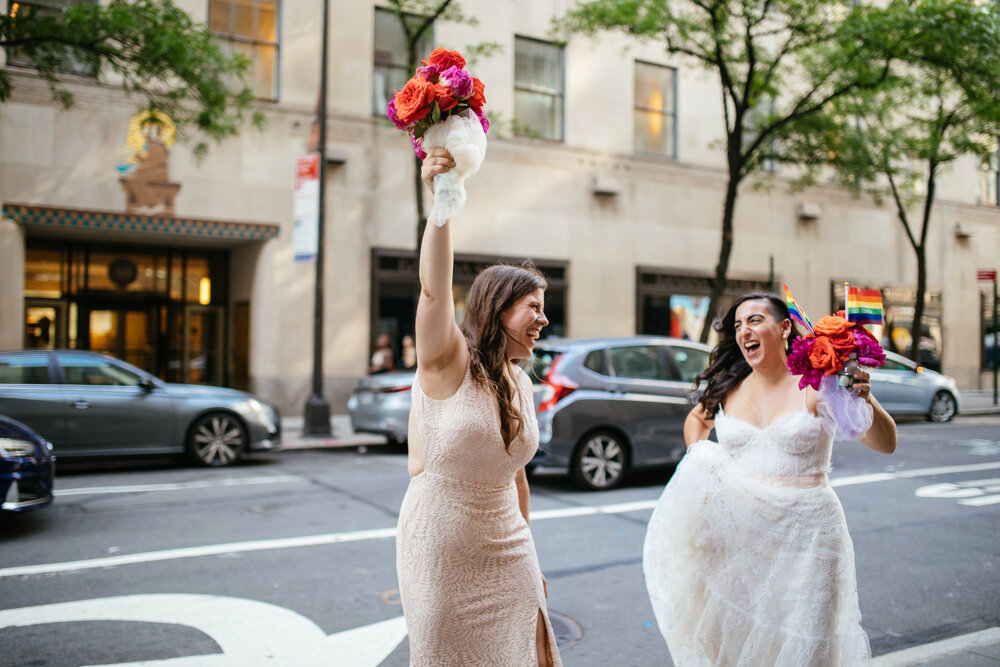 Two brides stand on the Manhattan sidewalk and hold their bouquets in the air while smiling at each other. One bride has two gay pride flags in her bouquet.

Luxury NYC Wedding Photography. Queer Wedding Photography. Inclusive Wedding Photographer. LGBTQ+ Manhattan Wedding.