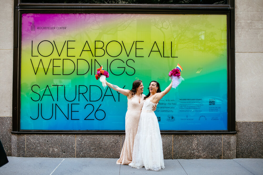 Two brides have arms around each other standing side by side and reaching their bouquets high up in the air, smiling at each other. They are standing on a Manhattan sidewalk in front of a massive sign that says "ROCKEFELLER CENTER / LOVE ABOVE ALL / WEDDINGS / SATURDAY / JUNE 26"

Luxury NYC Wedding Photography. Queer Wedding Photography. Inclusive Wedding Photographer. LGBTQ+ Manhattan Wedding.