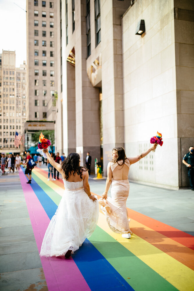 Two brides walk along the gay pride stripes painted on the ground outside Rockefeller Center. They are smiling at each other and holding their bouquets and gay pride flags in the air.

Luxury NYC Wedding Photography. Queer Wedding Photography. Inclusive Wedding Photographer. LGBTQ+ Manhattan Wedding.