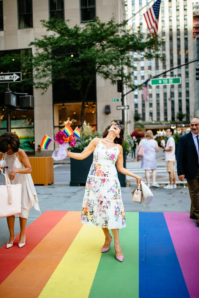 Woman in a floral dress stands outside Rockefeller Center and smiles up into the sky with her arms out. In one hand she has a purse and in her other hand she has a bouquet and three gay pride flags. The colors of the gay pride flag are painted in stripes on the ground below her feet.

Luxury NYC Wedding Photography. Queer Wedding Photography. Inclusive Wedding Photographer. LGBTQ+ Manhattan Wedding.