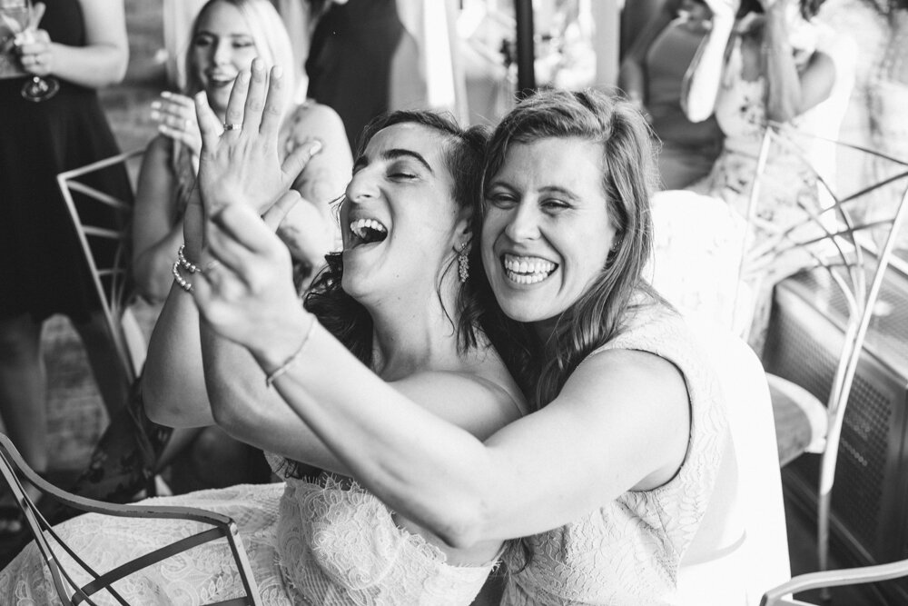 Bride holds the other bride's hand up in the air to display the wedding ring. They are both smiling big smiles and wedding guests look on and smile at them as well.

Luxury NYC Wedding Photography. Queer Wedding Photography. Inclusive Wedding Photographer. LGBTQ+ Manhattan Wedding.