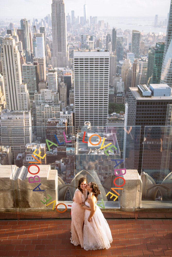 Two brides old each other in their arms and kiss on the cheek in front of a plexiglass wall that has "LOVE ABOVE ALL" painted in a circle on it. They are photographed from a floor above and the Manhattan skyline is visible behind them.

Luxury NYC Wedding Photography. Queer Wedding Photography. Inclusive Wedding Photographer. LGBTQ+ Manhattan Wedding.