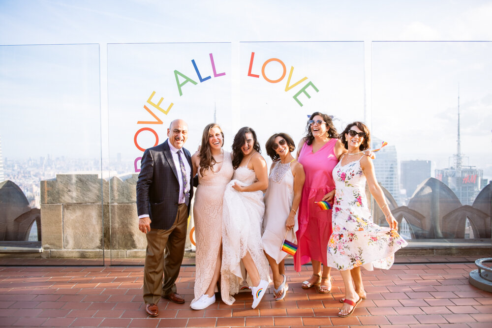 Brides and family stand next to each other at Top of the Rock and smile at the camera. They are posing in front of a plexiglass wall that has "LOVE ABOVE ALL" painted on it. The Manhattan skyline is behind them.

Luxury NYC Wedding Photography. Queer Wedding Photography. Inclusive Wedding Photographer. LGBTQ+ Manhattan Wedding.