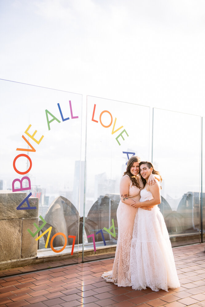Two brides have their arms around each other as they pose in front of a plexiglass wall with "LOVE ABOVE ALL" painted on it. The MAnhattan skyline is behind them.

Luxury NYC Wedding Photography. Queer Wedding Photography. Inclusive Wedding Photographer. LGBTQ+ Manhattan Wedding.