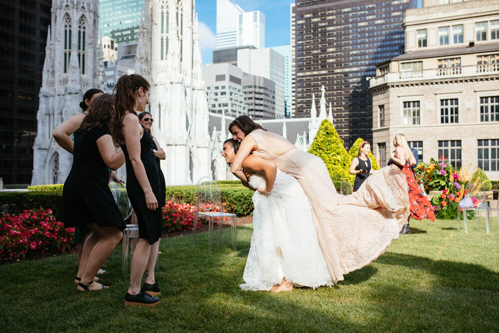One bride is leaning forward with the other bride behind her with her arms around her, lifted off of the ground. Wedding guests look at them and smile. They are at Top of the Rock and Manhattan buildings are visible behind them.

Luxury NYC Wedding Photography. Queer Wedding Photography. Inclusive Wedding Photographer. LGBTQ+ Manhattan Wedding.