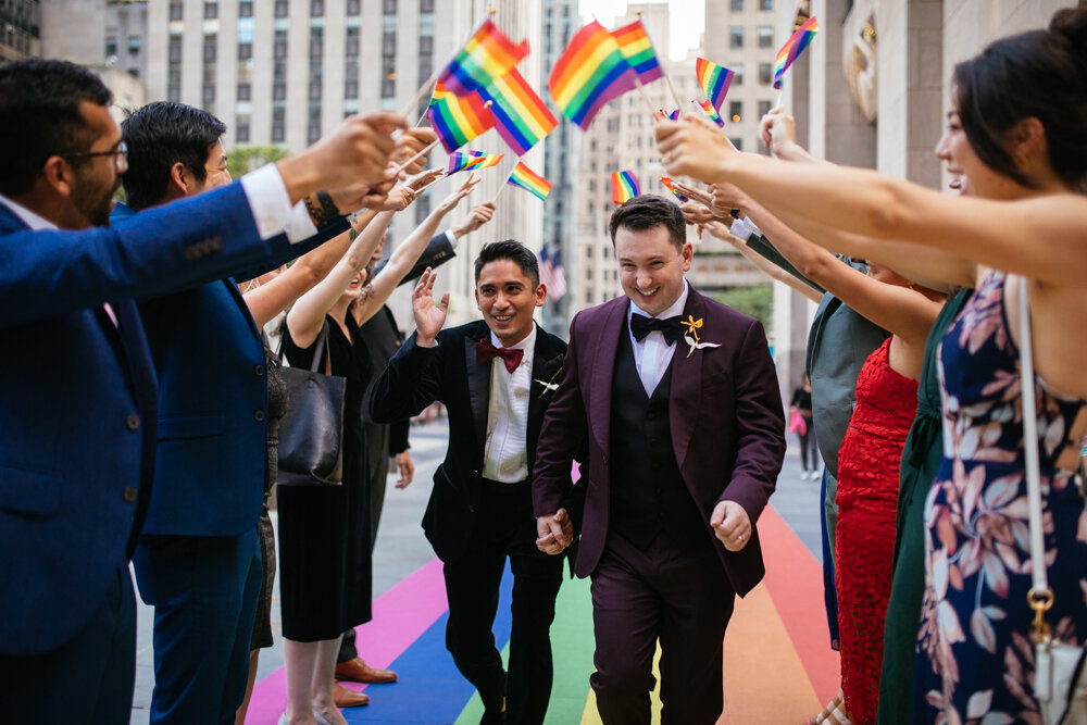 Grooms hold hands running along the gay pride flag painted on the ground outside Rockefeller Center. Their wedding guests are standing in two parallel lines reaching out with gay pride flags to create a tunnel for the grooms to run through.

Luxury NYC Wedding Photography. Queer Wedding Photography. Inclusive Wedding Photographer. LGBTQ+ Manhattan Wedding.