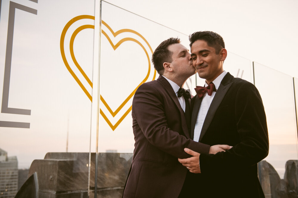 One groom kisses the other on the cheek as he smiles at the ground. Their arms are around each other and there is a heart painted on the plexiglass wall behind them at Top of the Rock.

Luxury NYC Wedding Photography. Queer Wedding Photography. Inclusive Wedding Photographer. LGBTQ+ Manhattan Wedding.
