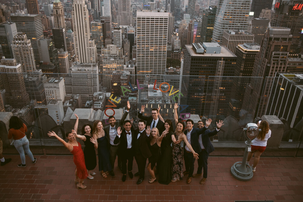Grooms and their wedding guests all pose with their arms in the air smiling at the camera, which is photographing them from a floor above. The Manhattan skyline is behind them.

Luxury NYC Wedding Photography. Queer Wedding Photography. Inclusive Wedding Photographer. LGBTQ+ Manhattan Wedding.