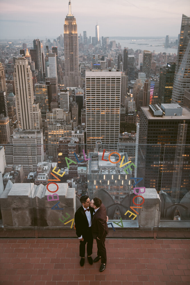 Grooms kiss at Top of the Rock in front of a painted "LOVE ABOVE ALL" on the plexiglass wall. They are photographed from a floor above and the Manhattan skyline is behind them.

Luxury NYC Wedding Photography. Queer Wedding Photography. Inclusive Wedding Photographer. LGBTQ+ Manhattan Wedding.