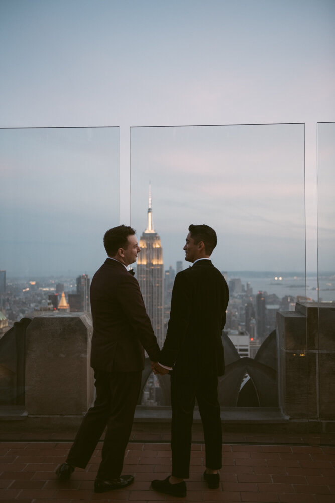 Grooms stand holding hands at the Top of the Rock and smiling at each other. The Manhattan skyline and Empire State Building are visible behind them.

Luxury NYC Wedding Photography. Queer Wedding Photography. Inclusive Wedding Photographer. LGBTQ+ Manhattan Wedding.