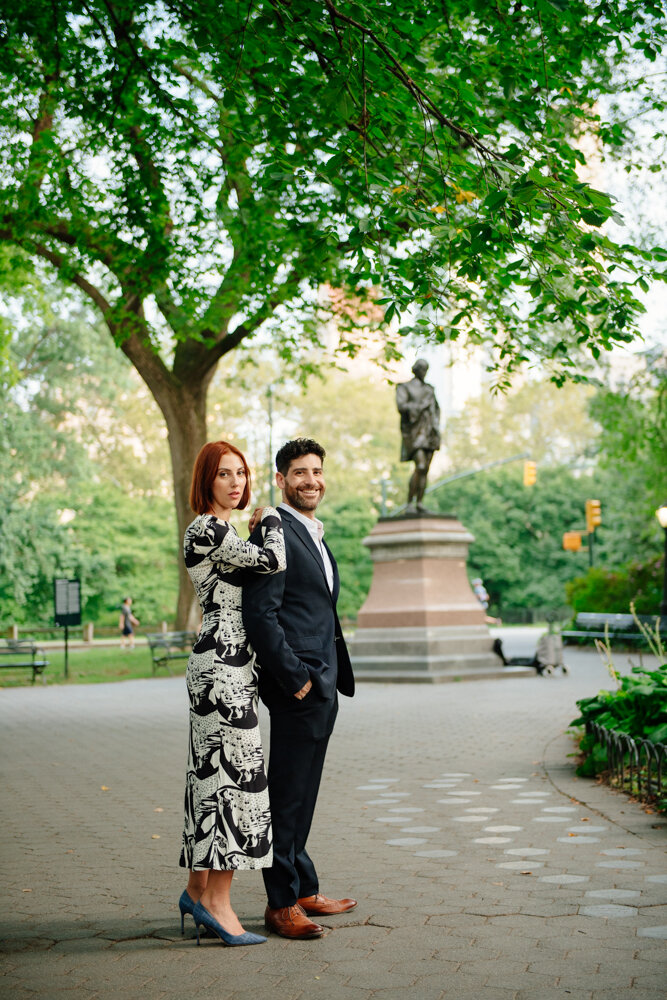 Woman stands behind man with her arms folded up on his shoulder. They are both looking at the camera and he is smiling. They are standing in Central Park and the trees and a statue are seen in the background.

Central Park Engagement Portraits. Manhattan Engagement Photography. NYC Engagement Photographer.
