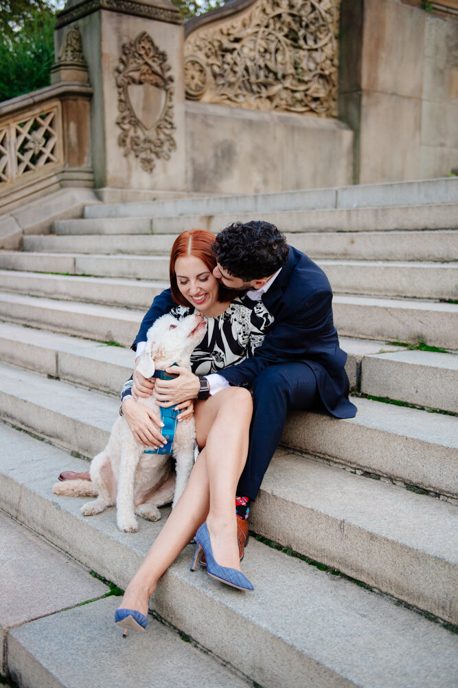 Man and woman and their dog sit on the steps in Central Park in Manhattan. The dog looks up at them as the woman looks down at the dog and the man kisses the woman on the cheek.

Central Park Engagement Portraits. Manhattan Engagement Photography. NYC Engagement Photographer.