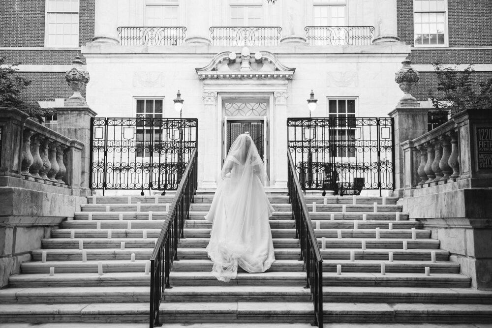 Bride holds the skirt of her wedding dress and ascends the steps of the Museum of the City of New York. The museum's entrance is seen in the background.

Luxury Local Wedding NYC. Wedding in Manhattan. New York City Wedding Photographer. Manhattan Luxury Wedding Photography. Museum of the City of New York Weddings.