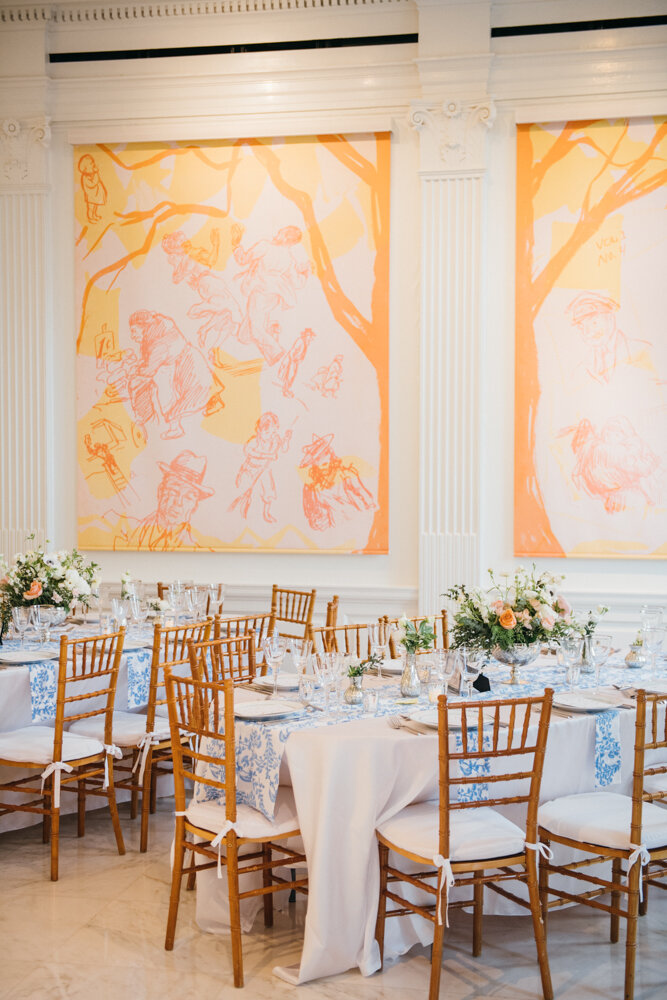 Tables are arranged with white and blue tablecloths and bouquets of flowers as centerpieces. Orange, pink, and yellow paintings on the wall are visible in the background.

Luxury Local Wedding NYC. Wedding in Manhattan. New York City Wedding Photographer. Manhattan Luxury Wedding Photography. Museum of the City of New York Weddings.