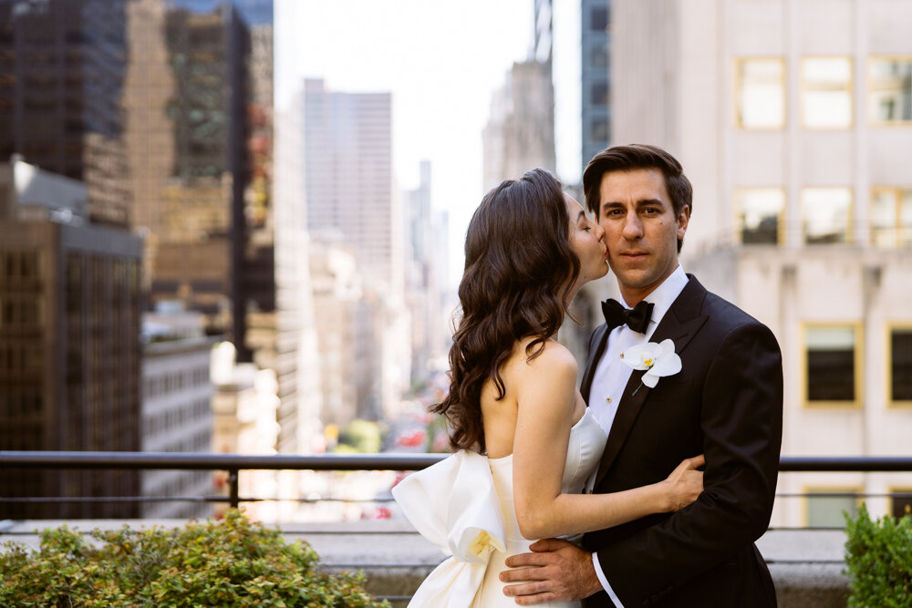 Groom looks at camera while the bride kisses him on the cheek. They have their arms around each other, standing on a Manhattan terrace.

University Club Wedding Photographer. Manhattan Luxury Wedding Photographer. Manhattan Bridal Portraits. Luxury Local Wedding NYC. 