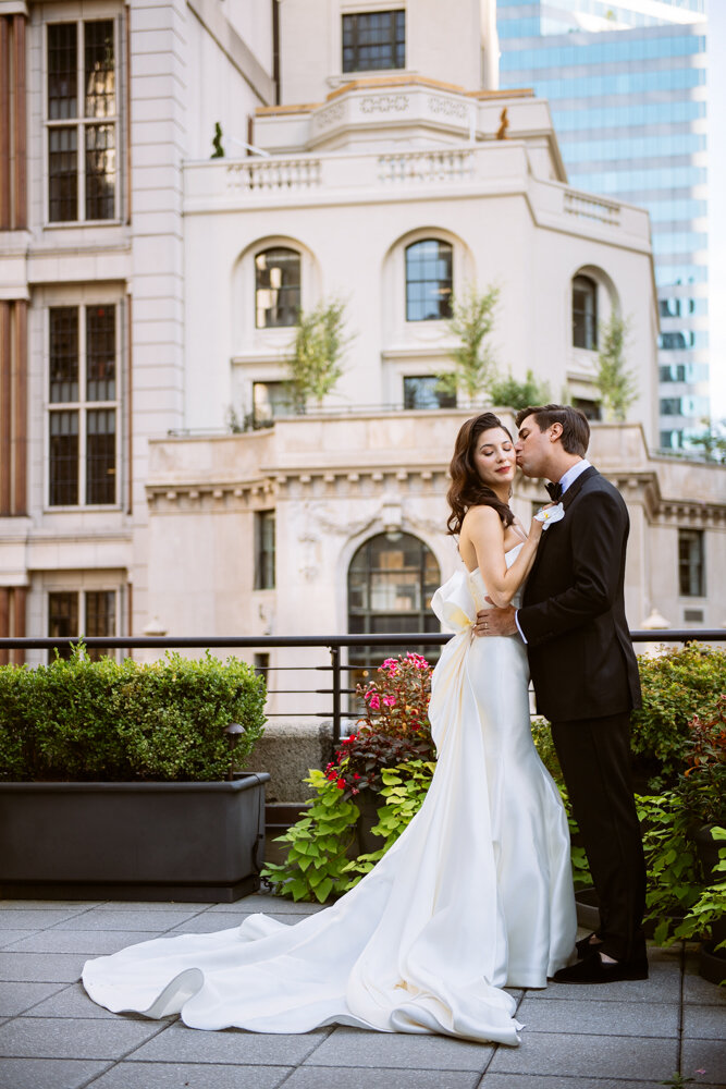 Groom kisses the bride on the cheek as they stand facing each other on a Manhattan terrace. The bride's wedding dress train extends behind her.

University Club Wedding Photographer. Manhattan Luxury Wedding Photographer. Manhattan Bridal Portraits. Luxury Local Wedding NYC. 