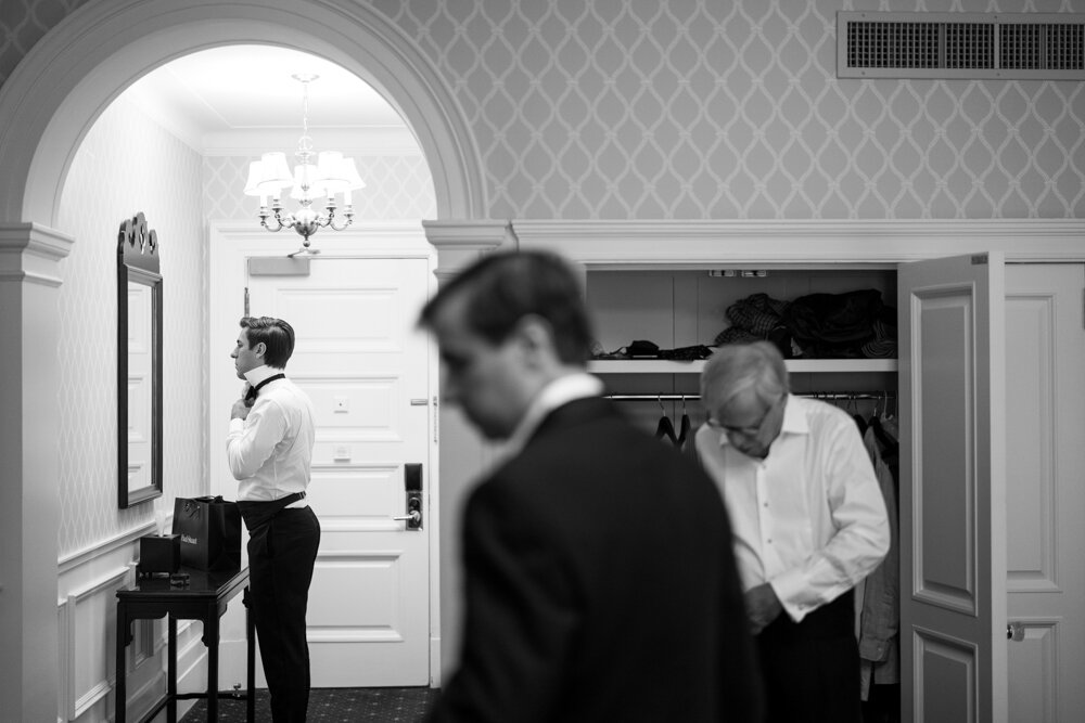 Groom is in focus in the background looking in the mirror and adjusting his tie. His father and groomsman are getting ready out of focus in the foreground.

University Club Wedding Photographer. Manhattan Luxury Wedding Photographer. Manhattan Groom Portraits. Luxury Local Wedding NYC. 