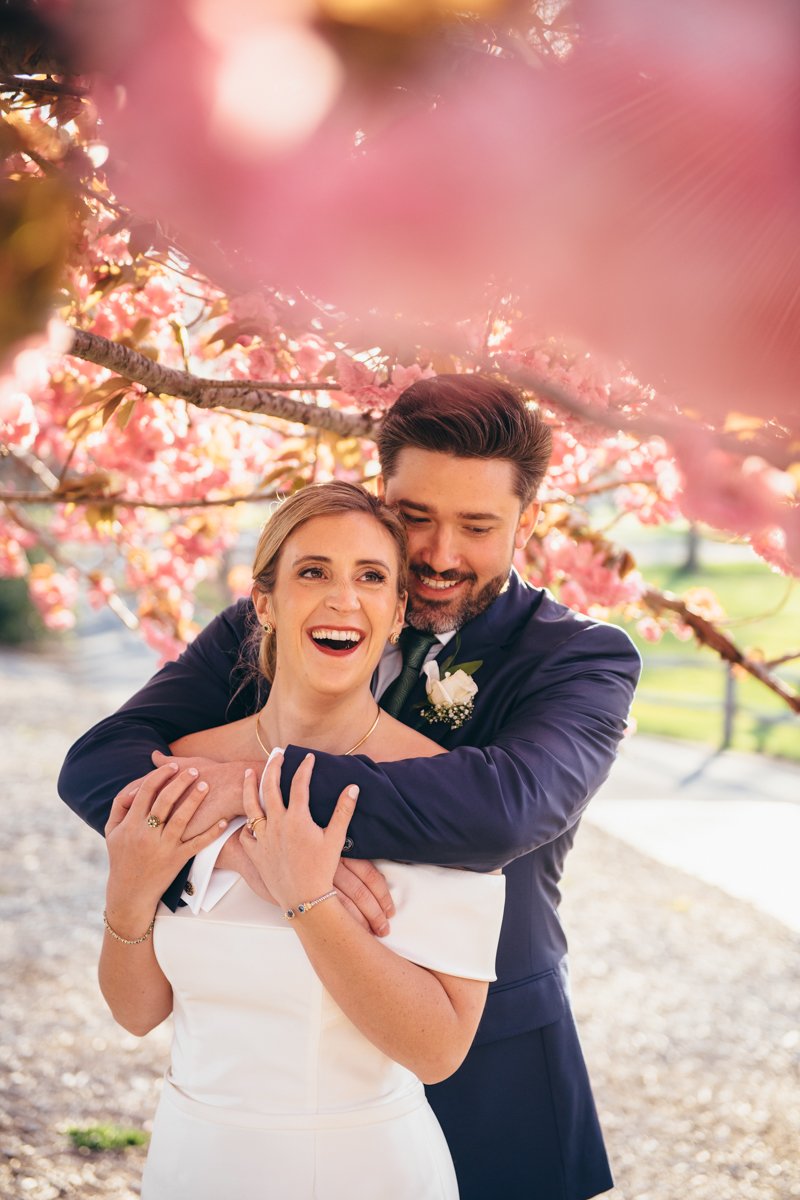 Groom stands behind the bride with his arms around her underneath a cherry blossom tree. Her hands reach up to hold his arms. They are both smiling off camera.

New York Wedding Photography. Long Island Wedding Photography. Luxury Local Wedding Photographer. Destination Wedding Photographer.