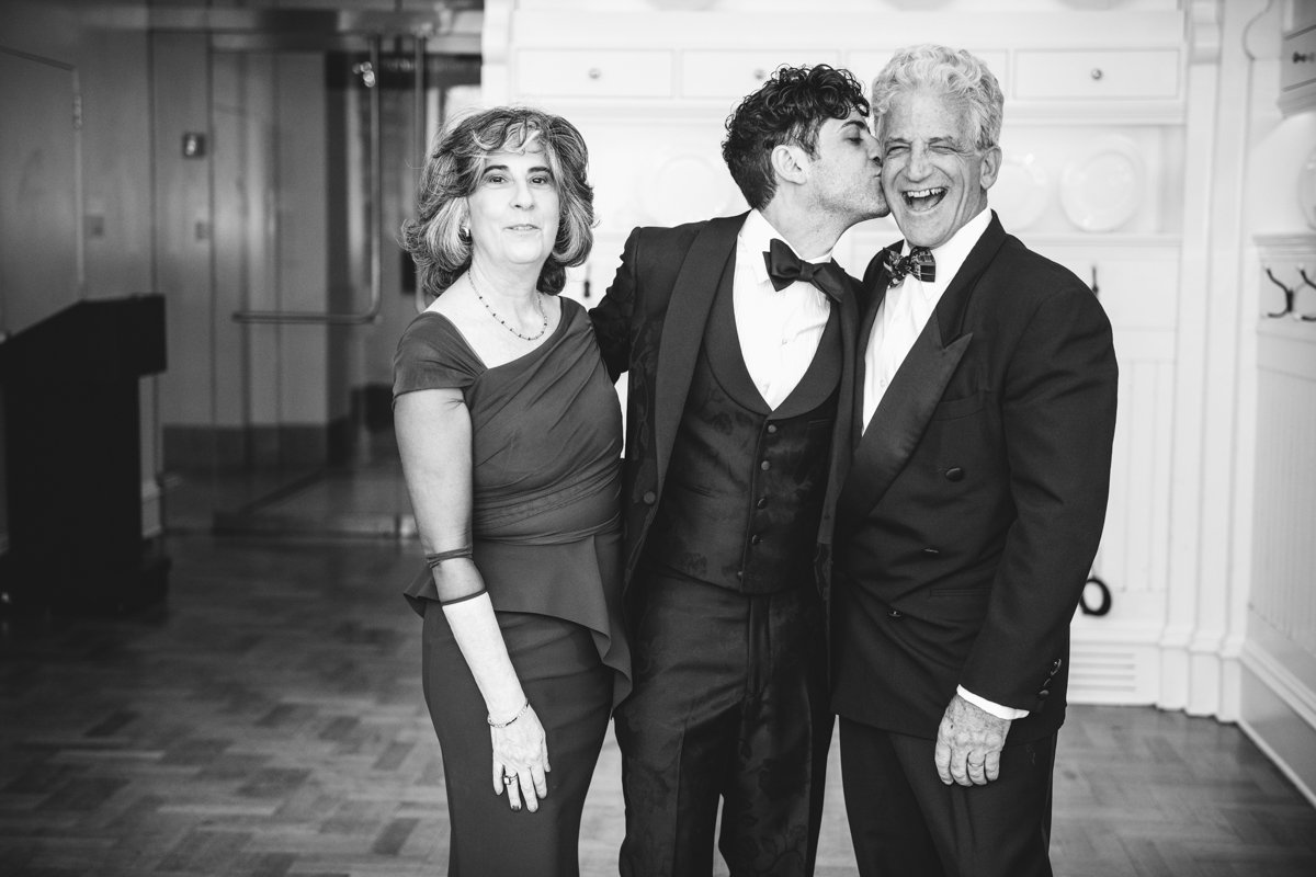 Groom has his arms around both parents and kisses his dad on the cheek.

Manhattan Wedding Photographer. New York Wedding Photographer. New York Historical Society Wedding. NY Historical Society Weddings.