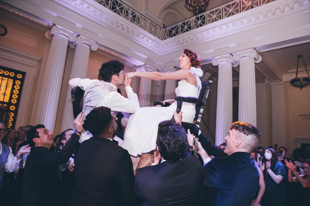 Bride and groom are seated in chairs that are being lifted up in the air by wedding guests. The groom is kissing the bride's hand.

Manhattan Wedding Photographer. New York Wedding Photographer. New York Historical Society Wedding. NY Historical Society Weddings.