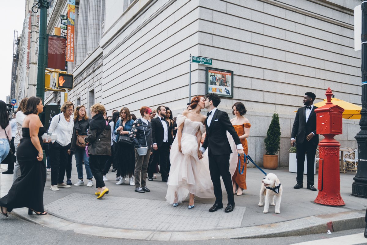 Bride and groom stand on a busy street corner and kiss. The groom is holding their dog's leash. Pedestrians pass by all around.

Manhattan Wedding Photographer. New York Wedding Photographer. New York Historical Society Wedding. NY Historical Society Weddings.