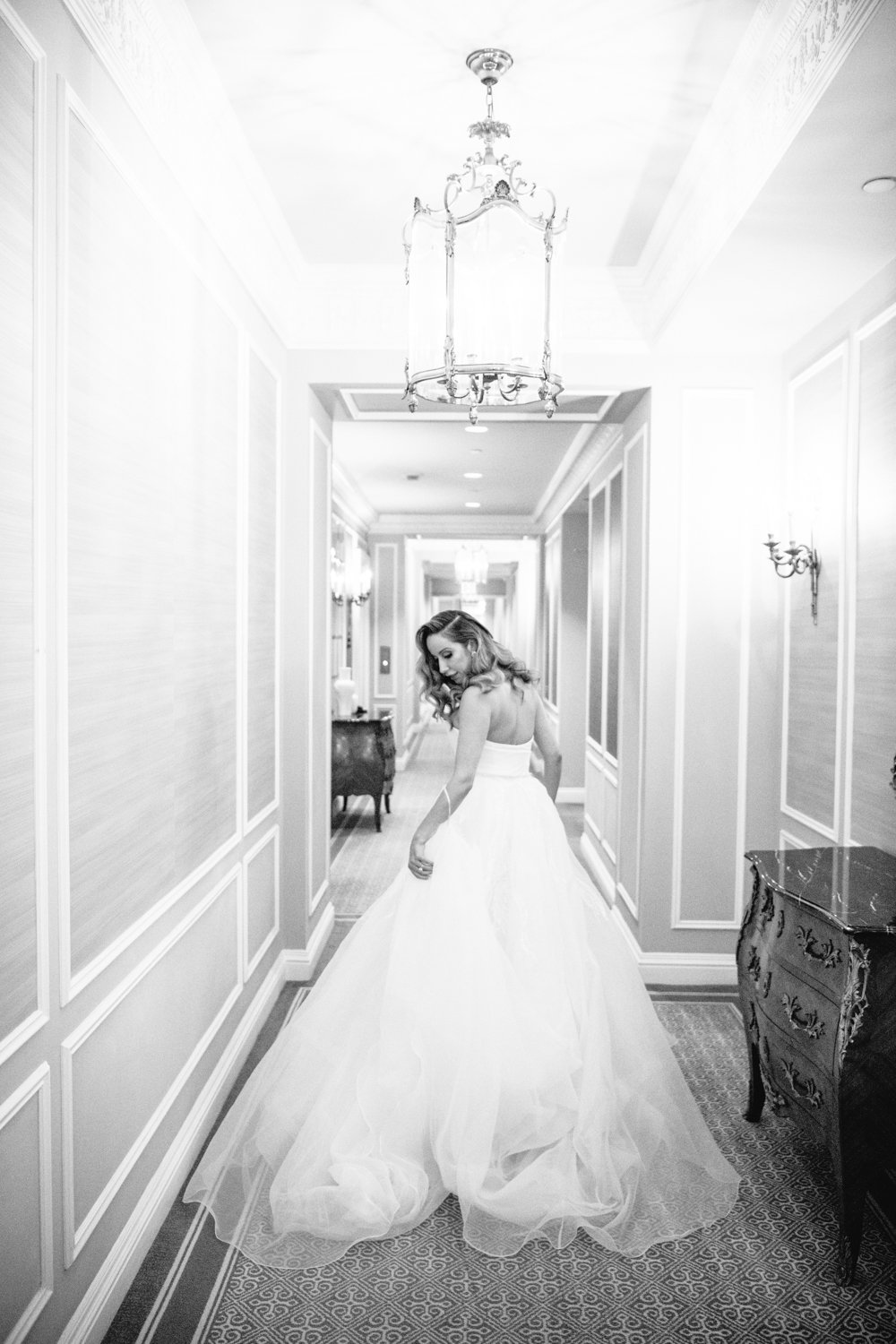 Bride is wearing a strapless wedding gown and walking away from the camera down the hallway of the St. Regis in Manhattan. She is looking back at the train of her dress.

Manhattan Luxury Wedding. New York Luxury Wedding Photographer. Wedding in Manhattan. NYC Luxury Wedding.