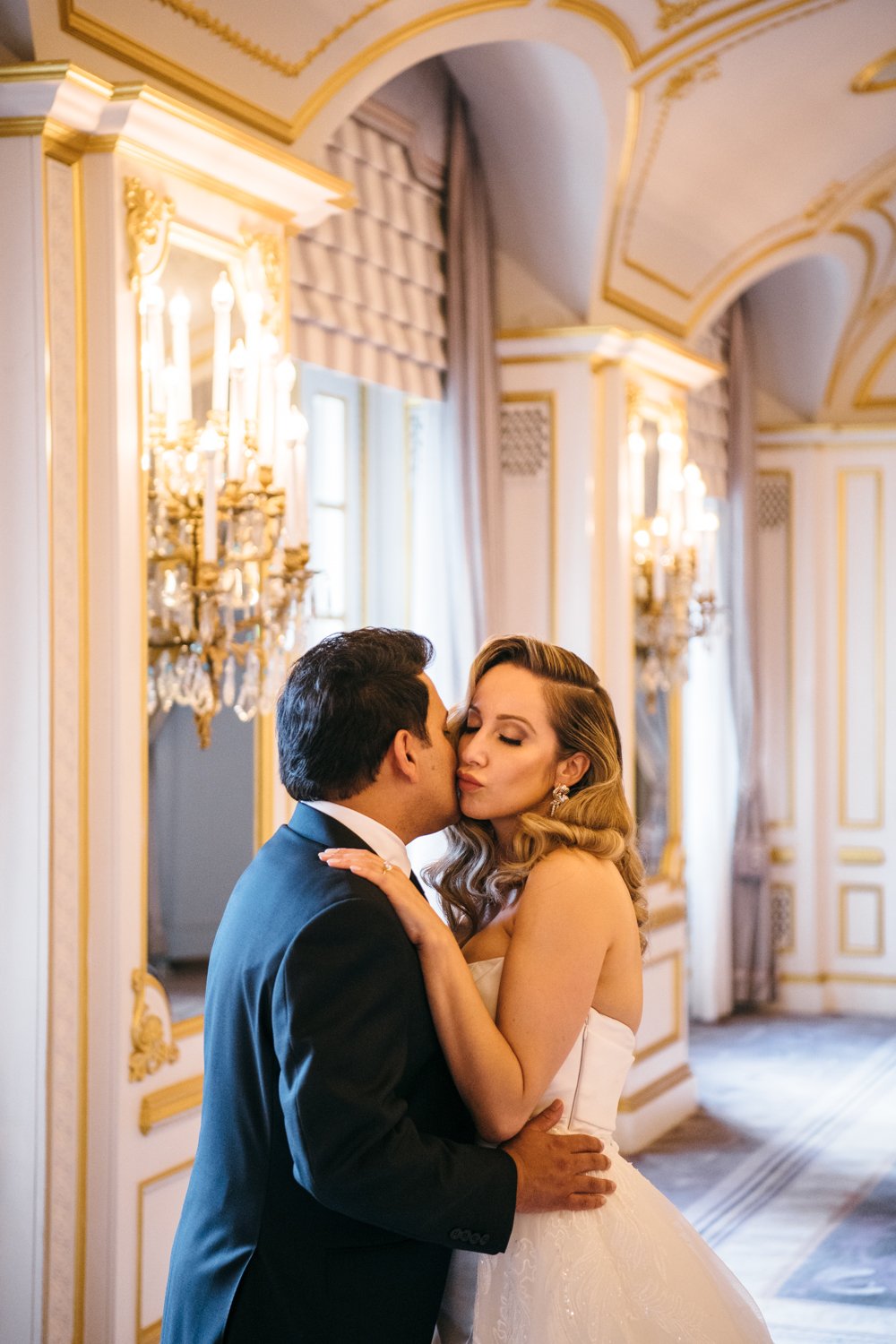 Bride and groom stand in the St. Regis in Manhattan. Groom has his hands around her waist and they kiss each other on the cheek.

Manhattan Luxury Wedding. New York Luxury Wedding Photographer. Wedding in Manhattan. NYC Luxury Wedding.