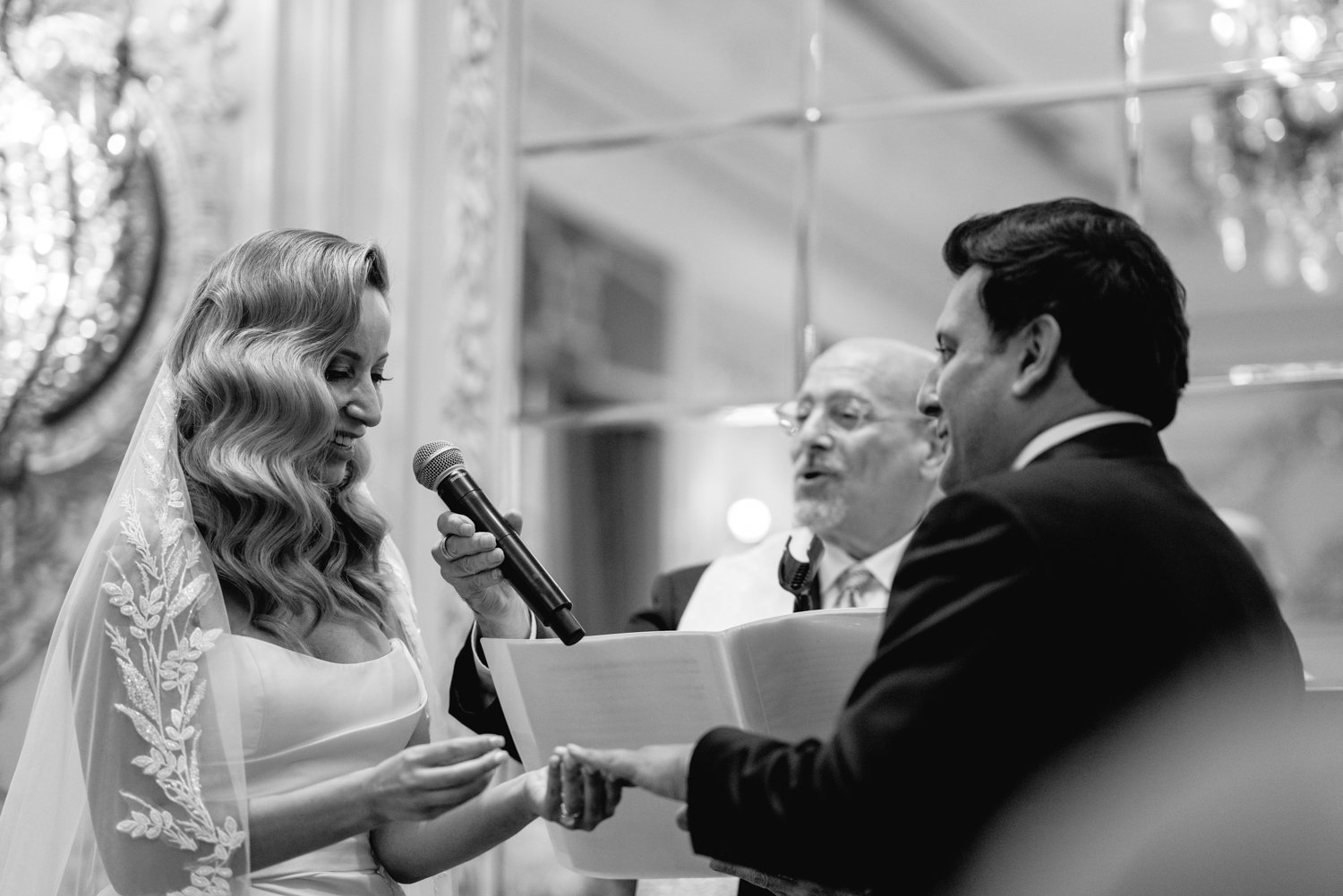 Bride holds the groom's hand and places a wedding ring on his finger. The officiant is behind them, holding a microphone up to the bride. They are all smiling. 

Manhattan Luxury Wedding. New York Luxury Wedding Photographer. Wedding in Manhattan. NYC Luxury Wedding.