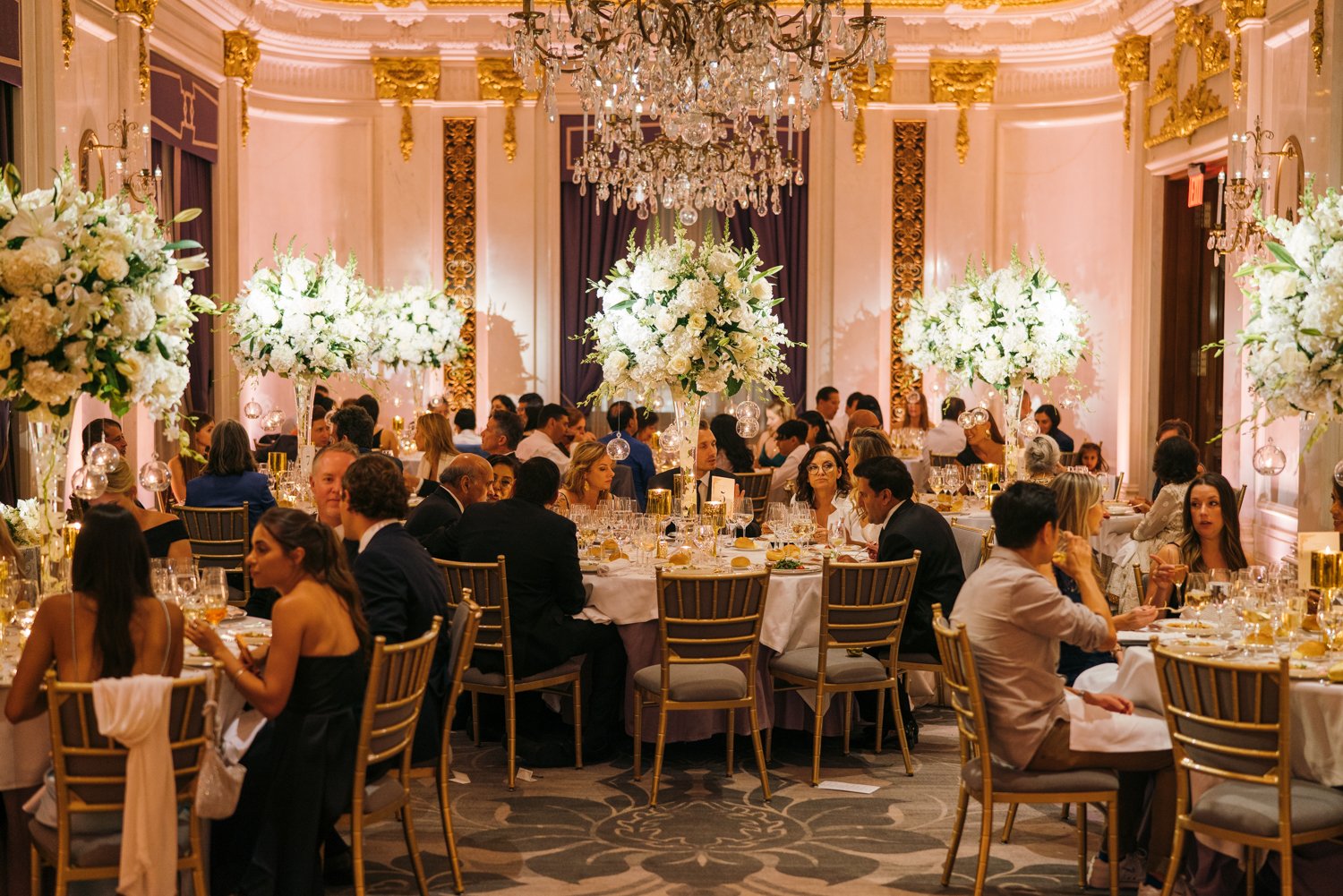 In the St. Regis in Manhattan, there are white floral bouquets above tables full of seated wedding guests.

Manhattan Luxury Wedding. New York Luxury Wedding Photographer. Wedding in Manhattan. NYC Luxury Wedding.