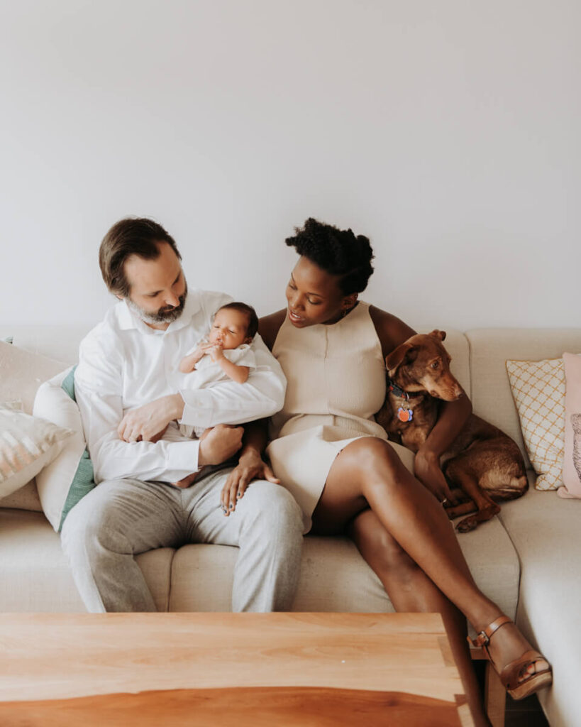 Mother and father sit next to each other on the couch and smile down at their baby. Mother has her arm around their dog.

Newborn Photography. Brooklyn Newborn Portraits. Lifestyle Newborn Photographer. Brooklyn Family Photography.