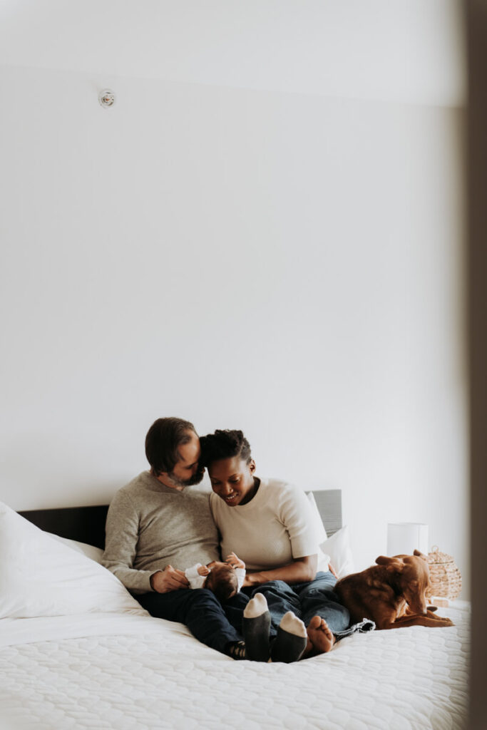 Mother and father sit in their bed next to their dog with their newborn baby in the father's lap as he kisses his wife on the cheek.

Newborn Photography. Brooklyn Newborn Portraits. Lifestyle Newborn Photographer. Brooklyn Family Photography.