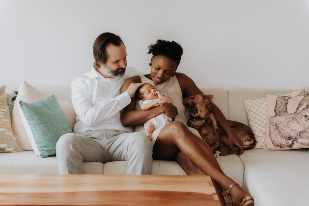 Mother and father sit on the couch with their dog next to them and their newborn baby in the mother's lap. They are both looking down at the newborn baby and smiling.

Newborn Photography. Brooklyn Newborn Portraits. Lifestyle Newborn Photographer. Brooklyn Family Photography.