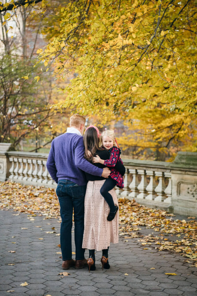 Husband and wife stand next to each other facing away from the camera. Woman has her daughter on her hip and her husband has his arm around her. Daughter is smiling at the camera. Photographed in Central Park.

Manhattan Family Portraits. Central Park Family Photography. NYC Family Photographer.