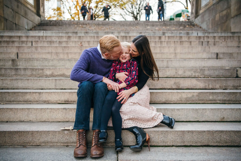 Husband, wife, and their daughter sit on steps in Central Park. The parents kiss their daughter on the cheek. Photographed in Central Park.

Manhattan Family Portraits. Central Park Family Photography. NYC Family Photographer.