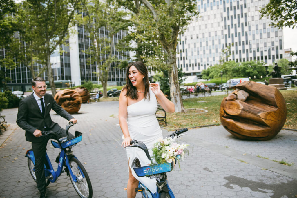 Woman is riding a citi bike in a white dress with flowers in the bike basket. A man in a suit is on a citi bike behind her. They are both smiling.

NYC Engagement Portraits. Manhattan Engagement Photographer. 
