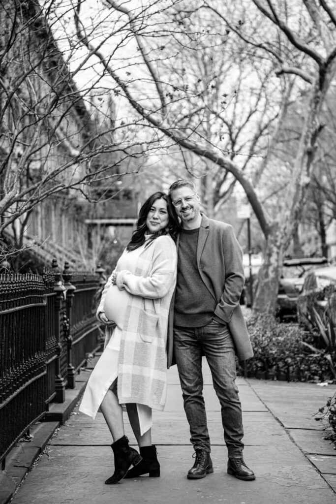 Husband and wife stand outside on a sidewalk in New York City. She has her hands on the baby hump and is leaning her back into her husband. They are both smiling at the camera.

Brooklyn Maternity Portraits. Brooklyn Family Photography. NYC Maternity Photographer.