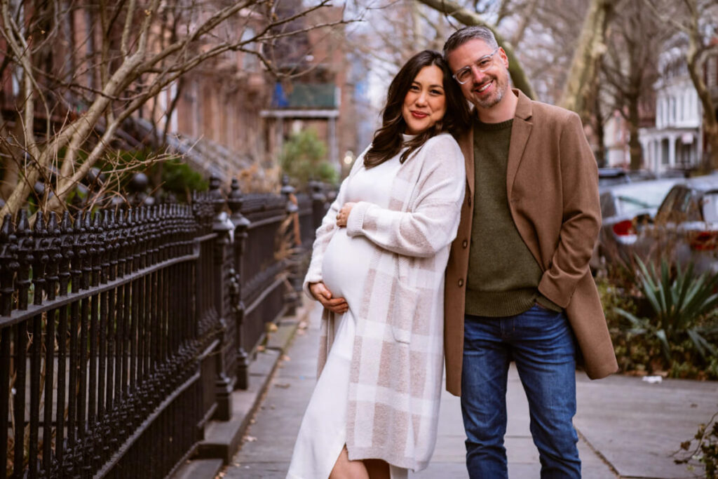 Husband and wife stand outside on a sidewalk in New York City. She has her hands on the baby hump and is leaning her back into her husband. They are both smiling at the camera.

Brooklyn Maternity Portraits. Brooklyn Family Photography. NYC Maternity Photographer.