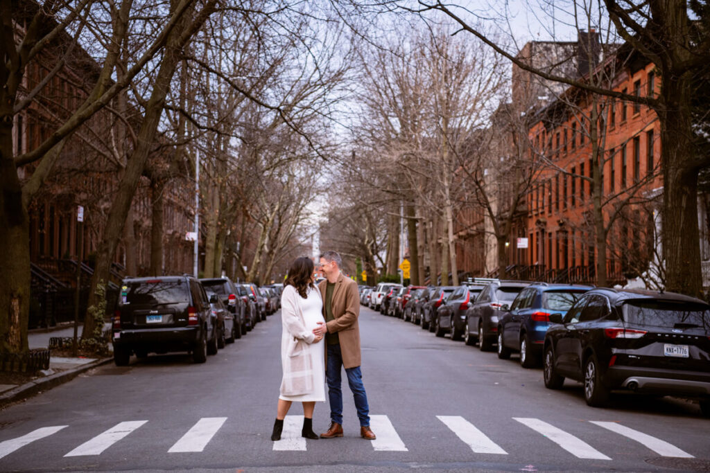 Husband and wife stand on a crosswalk in Brooklyn and both have a hand on her baby bump. They look at each other and smile. 

Brooklyn Maternity Portraits. Brooklyn Family Photography. NYC Maternity Photographer.