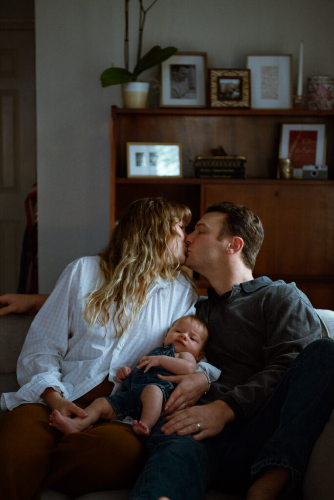 Husband and wife sit on the couch in their Austin, TX, home and kiss each other with their newborn baby in their lap.

Newborn Photography. Austin Newborn Photographer. Lifestyle Newborn Portraits. Austin Baby Portraits. Austin Famly Photography.