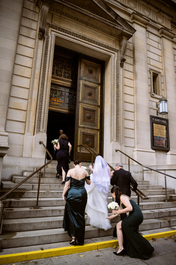 Bride is walking up the steps of the Church of St. Ignatius Loyola with her parents and bridesmaids helping her up the stairs.

Luxury NYC Wedding Photography. Manhattan Luxury Wedding Photographer. NYC Luxury Wedding. University Club Wedding Photographer.