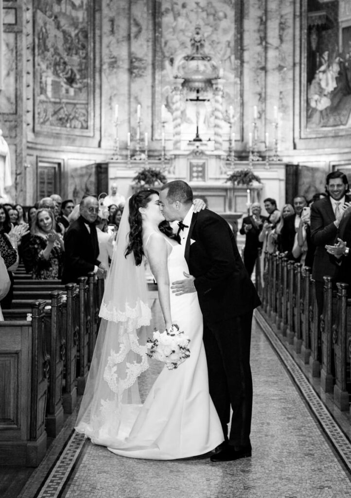 Bride and groom kiss each other at the top of the aisle in the church. Wedding guests look on, smiling and clapping. Black and white photograph.

Luxury NYC Wedding Photography. Manhattan Luxury Wedding Photographer. NYC Luxury Wedding. University Club Wedding Photographer.