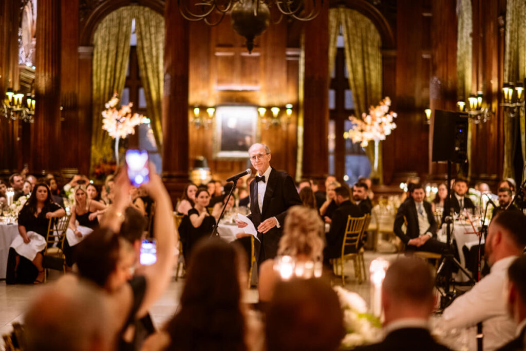 The father of the bride is standing at the center of the room with a microphone on a stand in front of him in the University Club in Manhattan. Wedding guests are seated at tables all around him. 

Luxury NYC Wedding Photography. Manhattan Luxury Wedding Photographer. NYC Luxury Wedding. University Club Wedding Photographer.