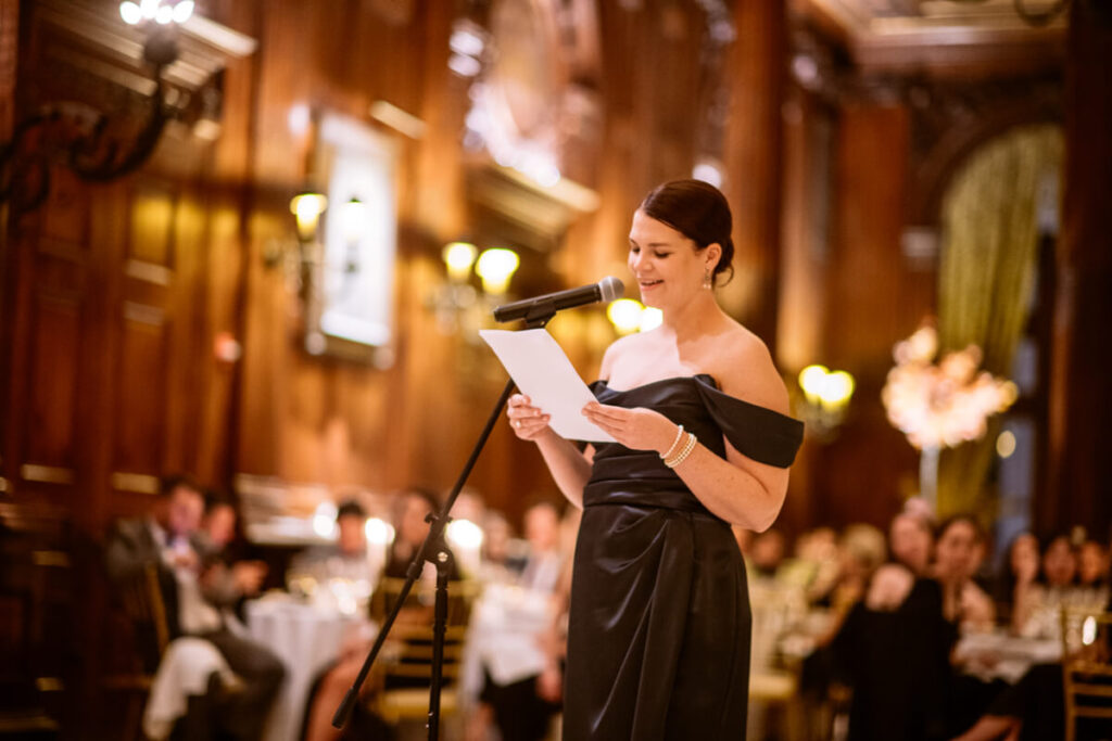 Maid of honor is wearing an off the shoulder navy dress, reading from a paper into a microphone. Wedding guests are in the background, seated at tables in the University Club in Manhattan.

Luxury NYC Wedding Photography. Manhattan Luxury Wedding Photographer. NYC Luxury Wedding. University Club Wedding Photographer.